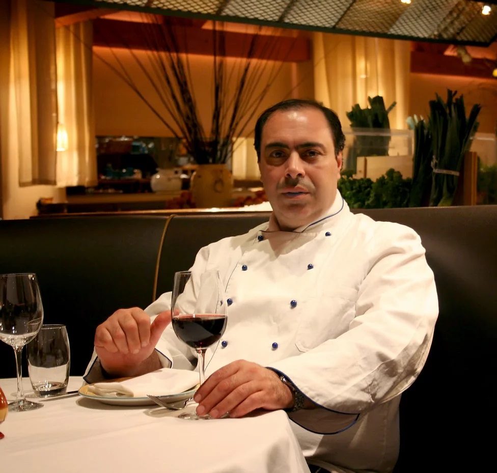 Chef Zapantis sitting at a table in a white coat with a glass red wine.